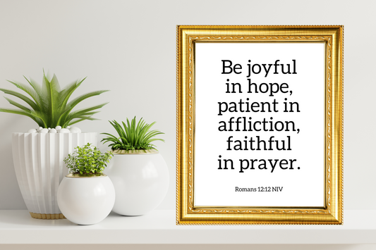 High-definition decorative inspirational wall art printable of Romans 12:12 styled in a gold frame, surrounded by potted plants on a white surface.