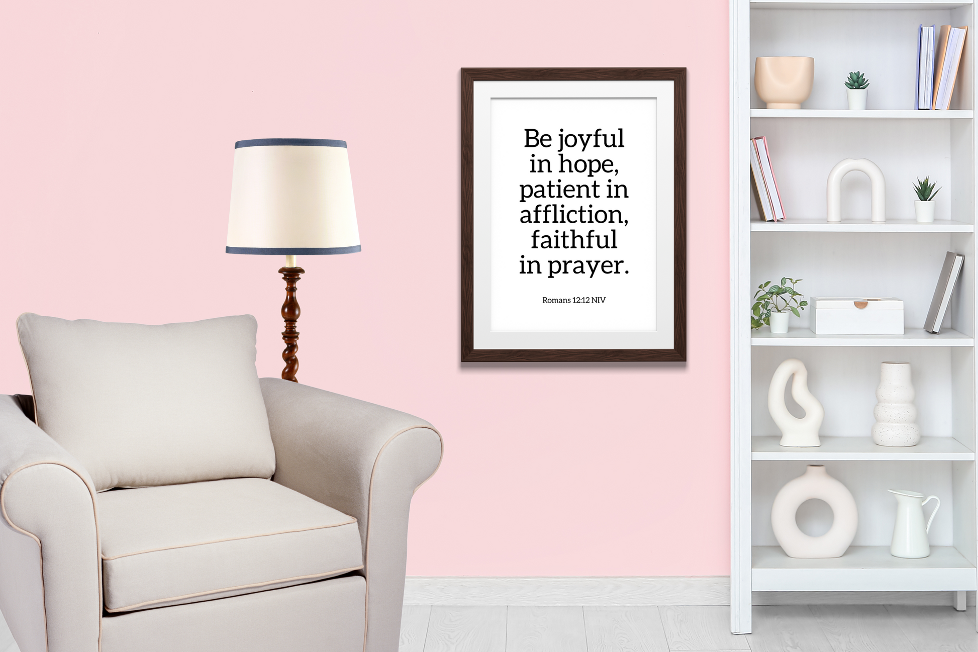 Contemporary printable wall art featuring Romans 12:12 bible verse in a frame hanging up in a living room with a framed quote on a pink wall, a lamp, and a bookshelf.