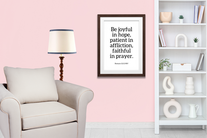 Contemporary printable wall art featuring Romans 12:12 bible verse in a frame hanging up in a living room with a framed quote on a pink wall, a lamp, and a bookshelf.