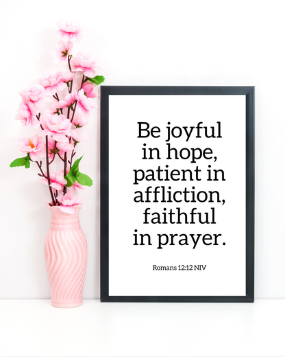 Framed quote 'Be joyful in hope, patient in affliction, faithful in prayer' with pink vase and flowers on white background