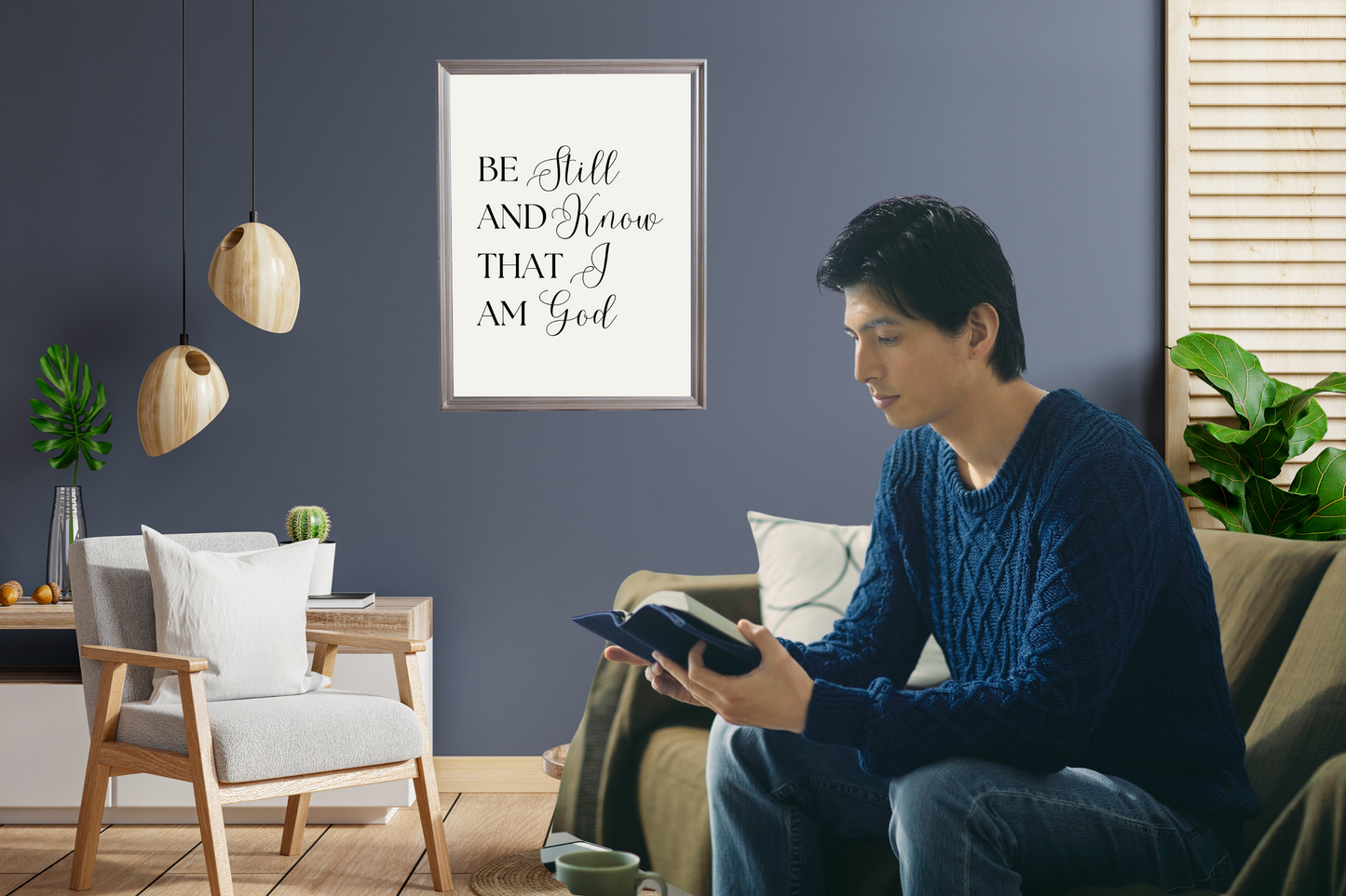 Man sitting on a couch reading a book in a living room with motivational wall art 'Be still and know that I am God.'