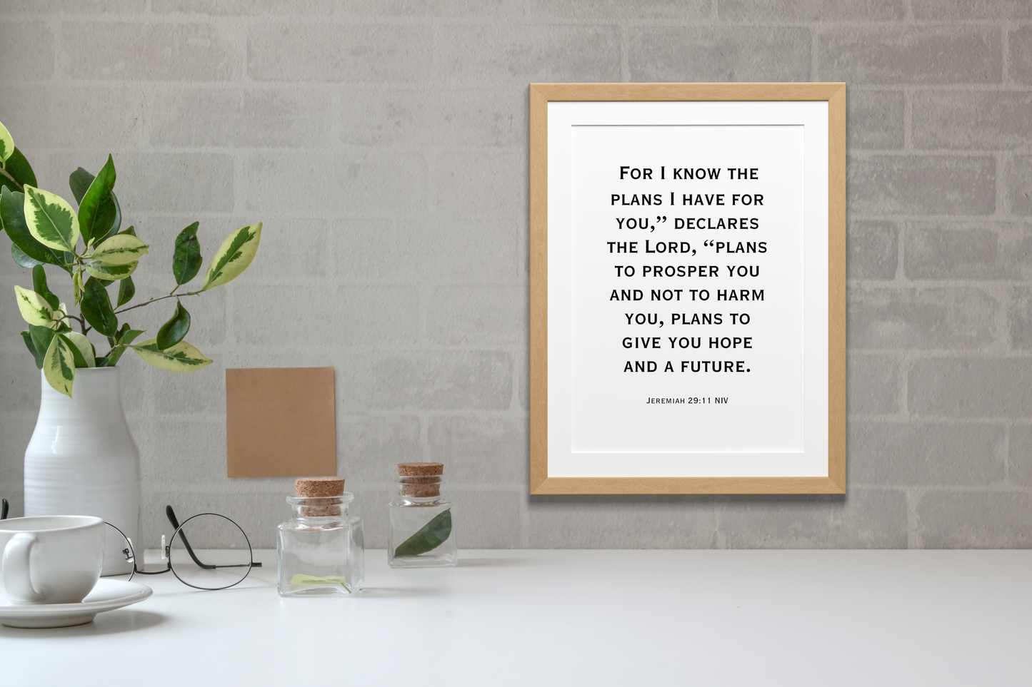 Contemporary printable wall art featuring Jeremiah 29:11 bible verse in clean black typography with a wooden frame on a brick wall background, above a table with a cup and plants.